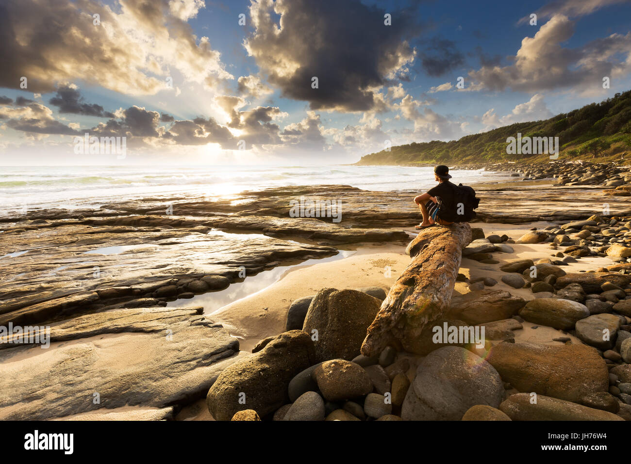 A person sits on a beautiful rocky beach and watches as the sun bursts through the clouds over the sea in this beautiful seascape. Stock Photo