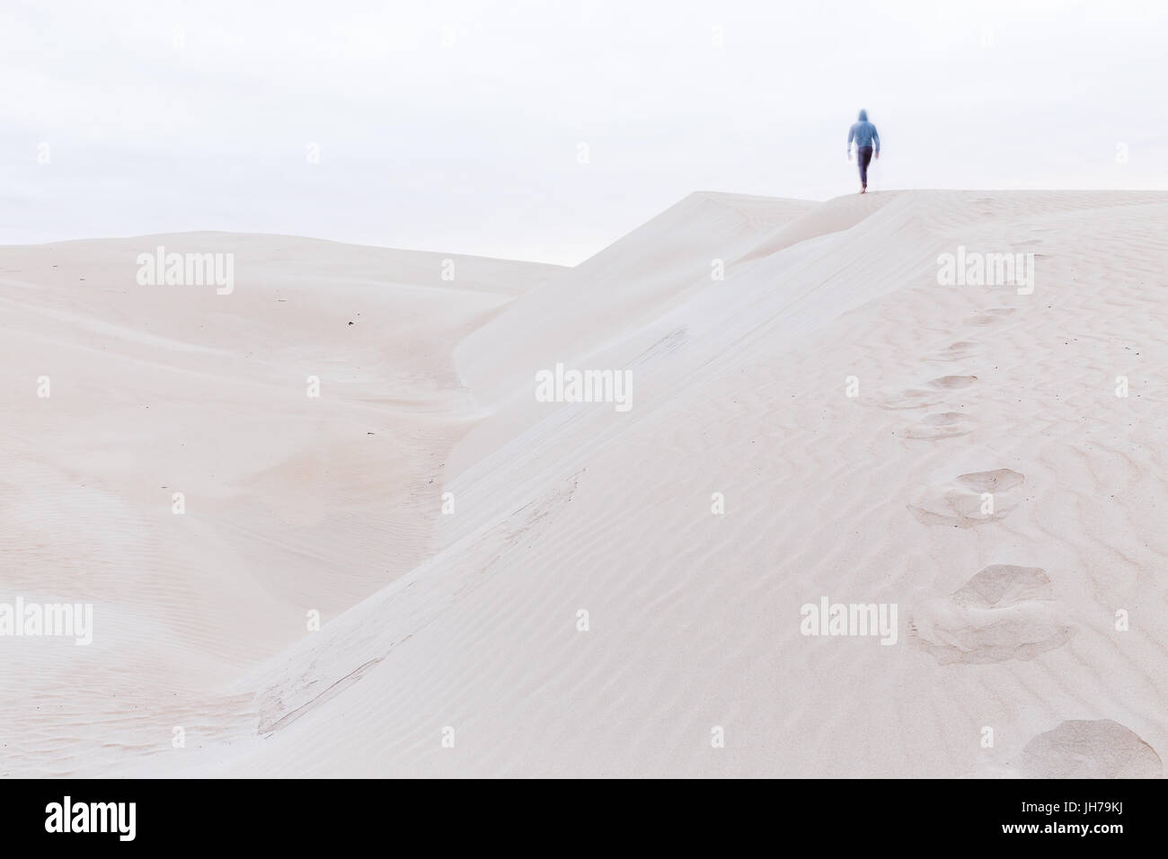 A person walks along a sand dune ridge leaving behind a trail of footprints in the sand. Stock Photo