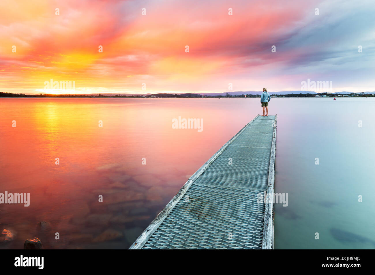 A person watches a beautiful, colourful sunset over the sea from the end of a small jetty. Stock Photo