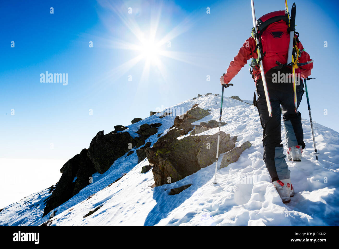 Mountaineer walking up along a steep snowy ridge with the skis in the backpack. In background a dramatic sky with a shiny bright sun. Concepts: advent Stock Photo