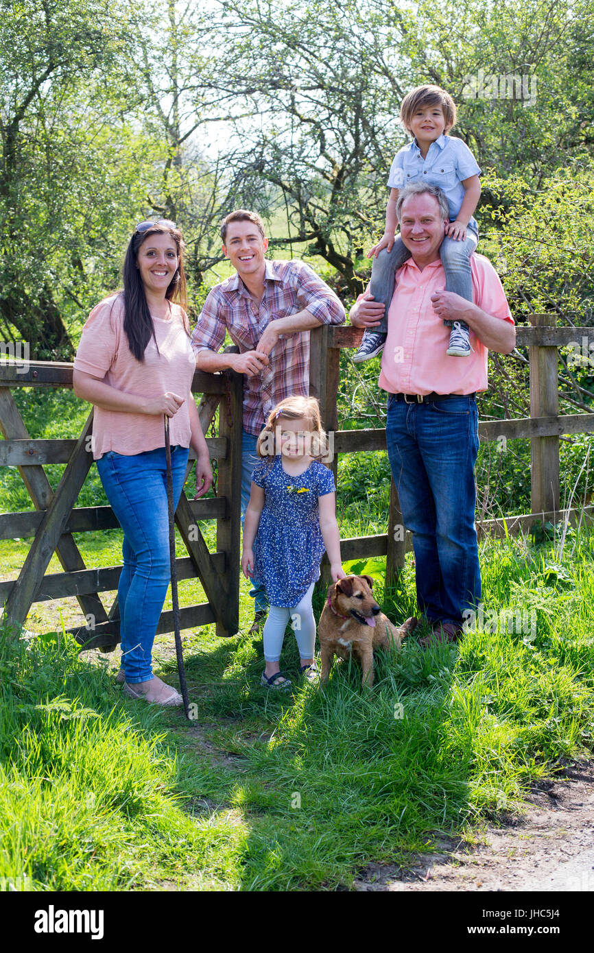 Family posing for the camera at a farm gate in the countryside. There are two children, their parents, a grandfather and a pet dog. The little boy is  Stock Photo