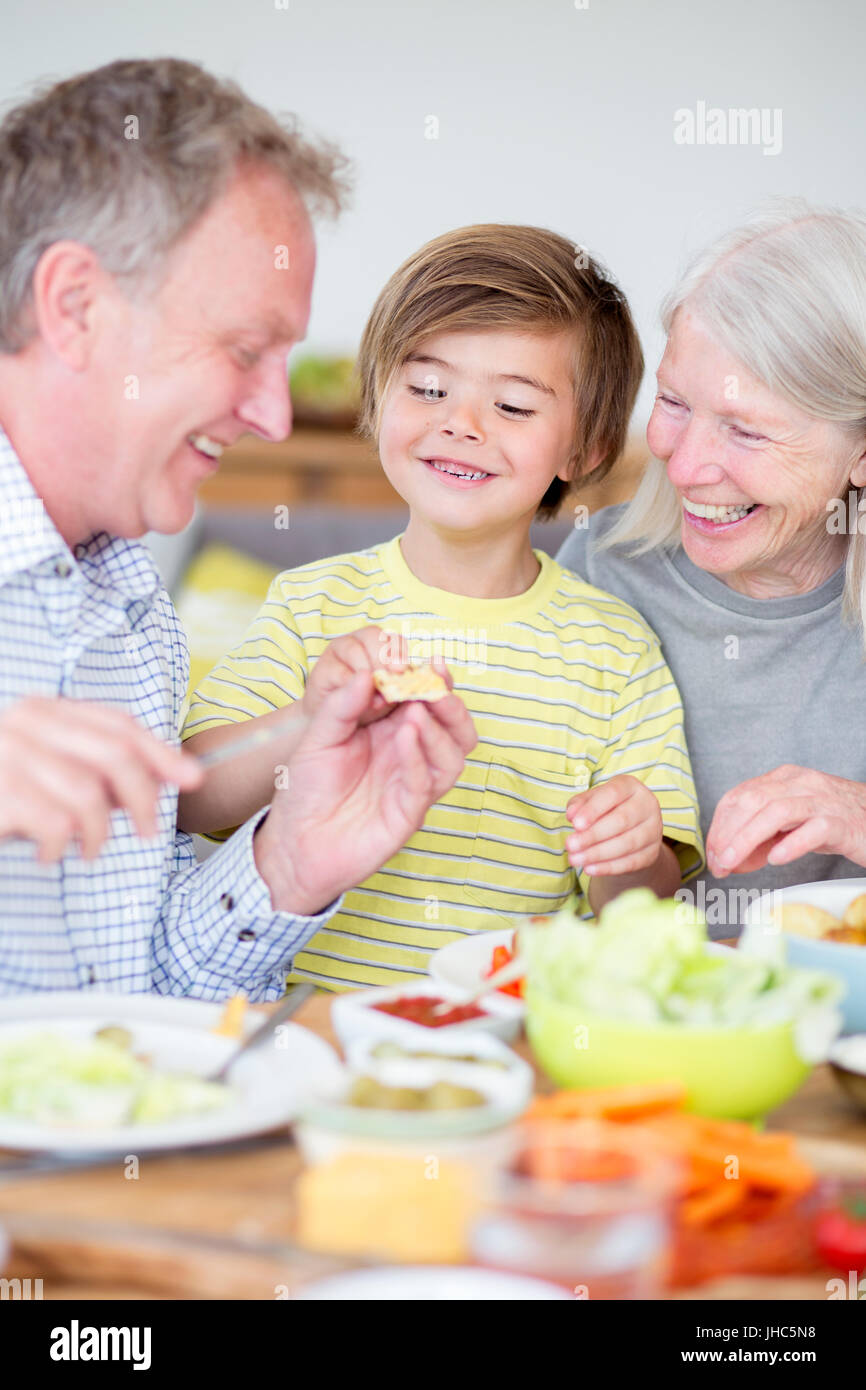 Little boy is sitting at the dining table with his grandparents in their home. His grandfather is buttering some bread and giving it to his grandson. Stock Photo