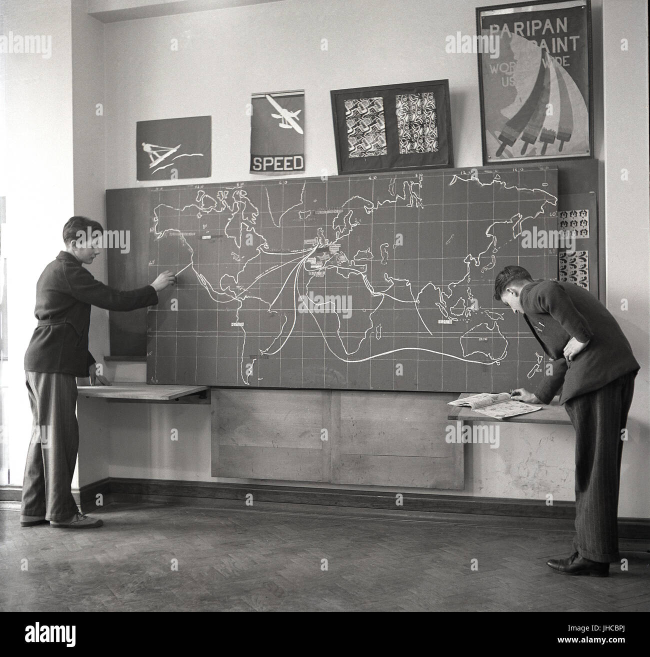 1950s, historical, a male student points to the route from America to the UK on a large blackboard, which has a map of the world showing the different trading routes products such as cod liver oil and honey - and even dried blood! - take to reach their markets. Stock Photo