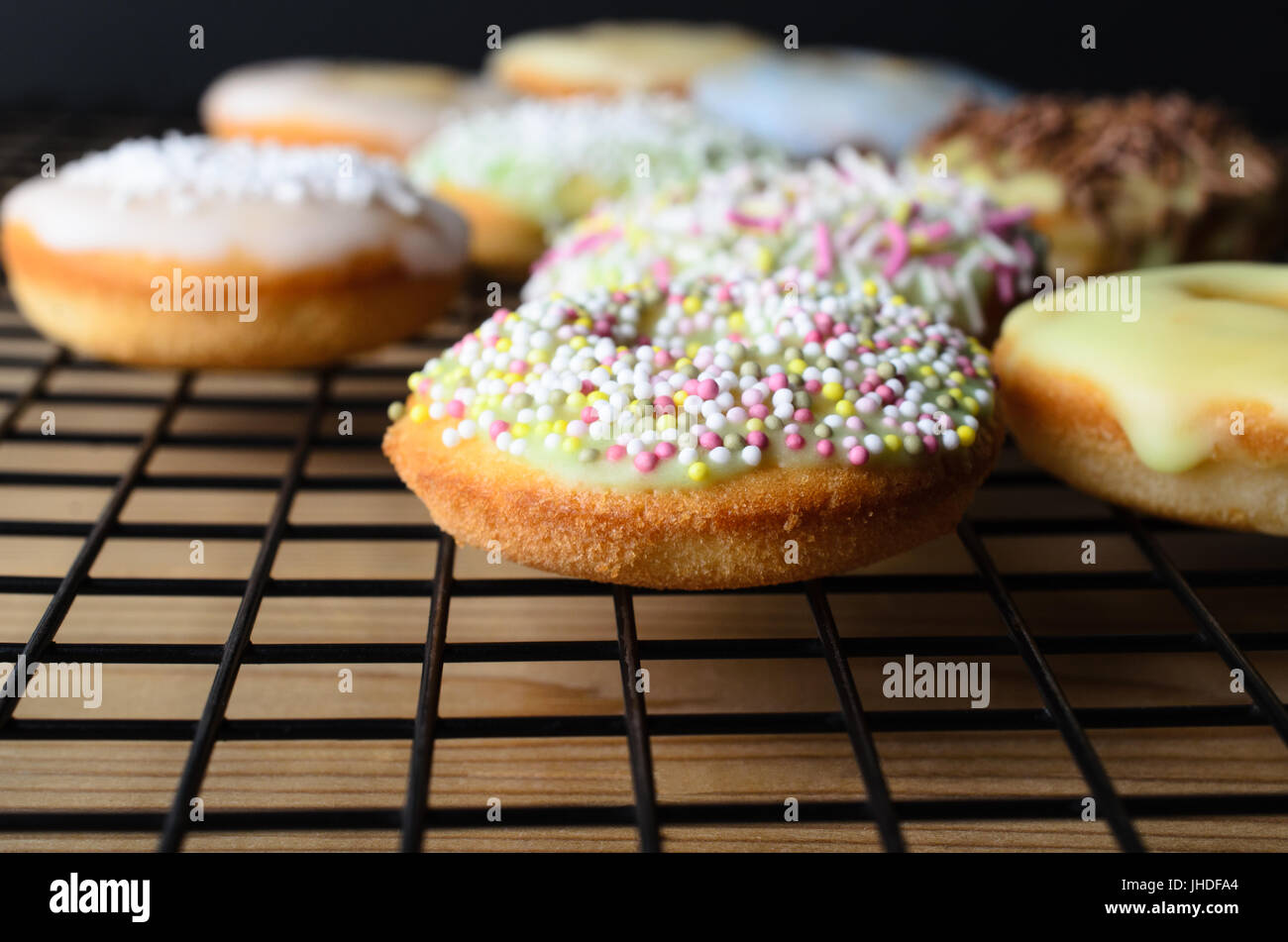 Home baked mini doughnut sponge cakes, with a variety of decorative sprinkles, on a black wire cooling rack and wooden table. Stock Photo