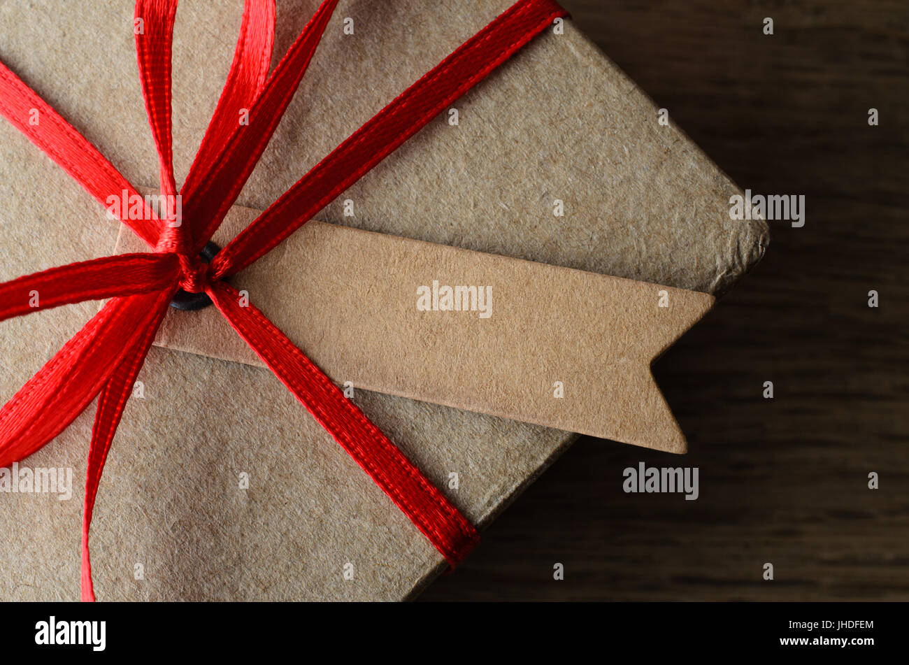 Overhead close up of a blank label tied to a plain brown  gift box with red ribbon on oak wood surface. Stock Photo