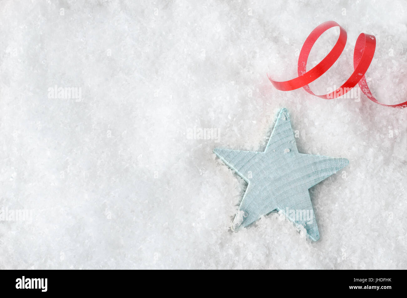 Overhead Christmas background shot.  A pale blue wooden star, resting in white artificial snow, with red swirl of ribbon above and copy space to the l Stock Photo