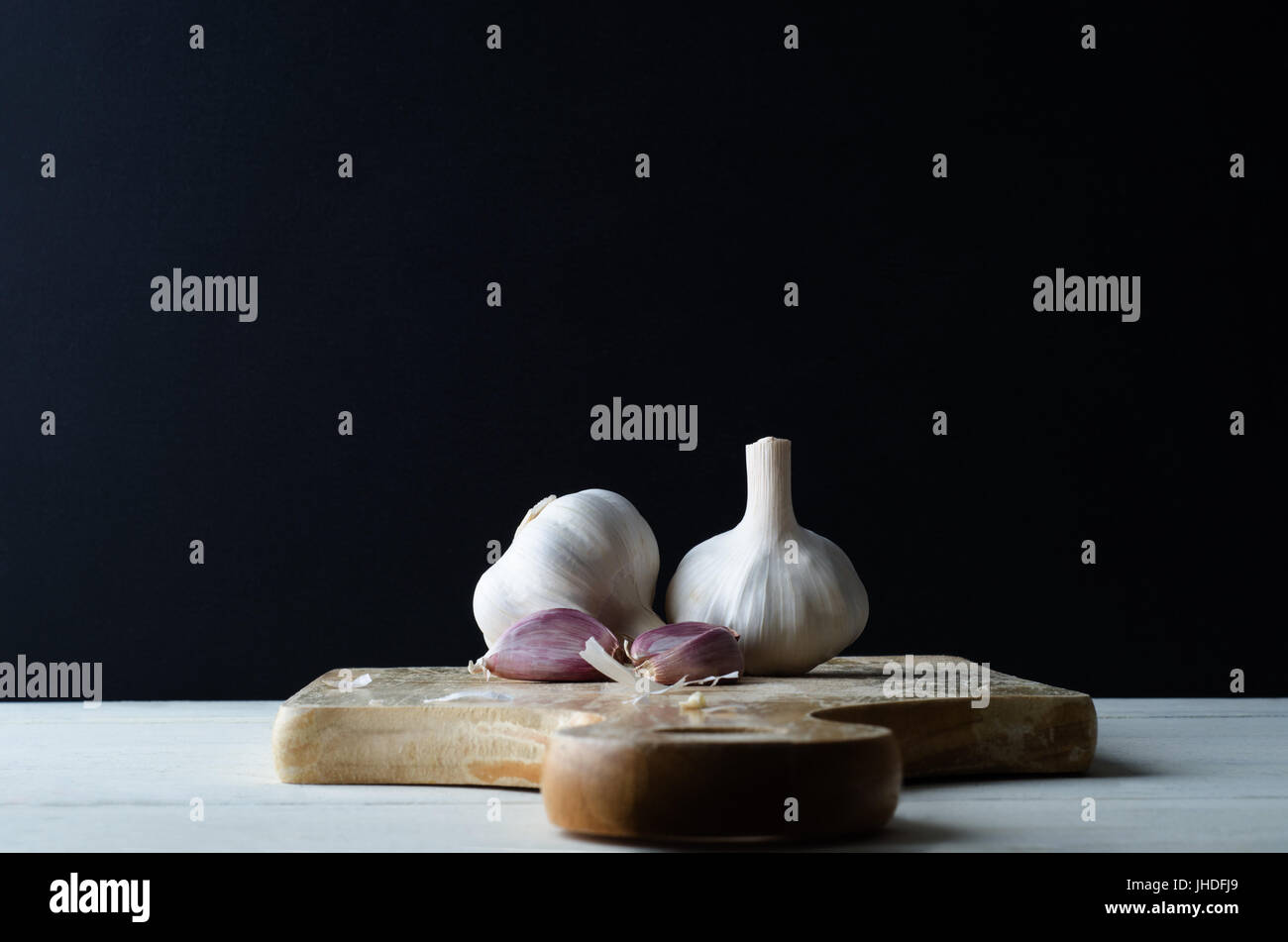 Food preparation still life of two garlic bulbs and cloves with scattered peelings on an old wooden chopping board with white plank kitchen table bene Stock Photo