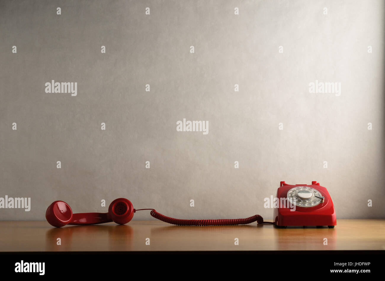Eye level shot of a retro red dial up telephone on a light wood veneer desk or table with handset taken off the hook and trailed across it.  Off white Stock Photo