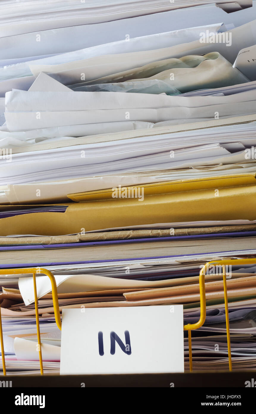 A wire office tray labeled 'In', piled high with papers, documents and folders that reach to the top of the frame and extending beyond it. Stock Photo