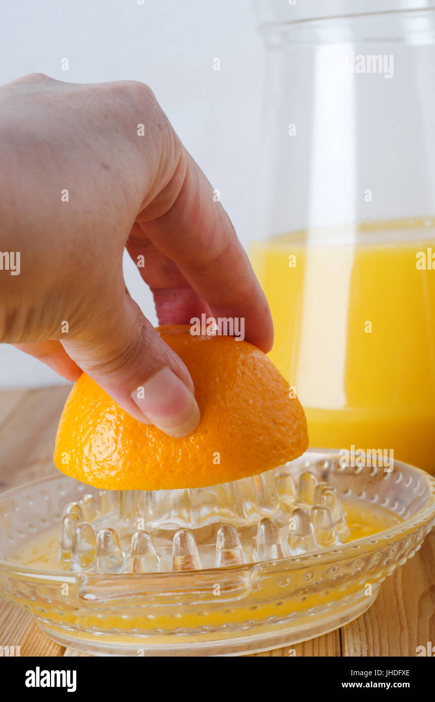 Hand reaching into frame from left, squeezing half an orange on glass manual fruit squeezer.  Jug of fresh orange juice in background on light wood pl Stock Photo