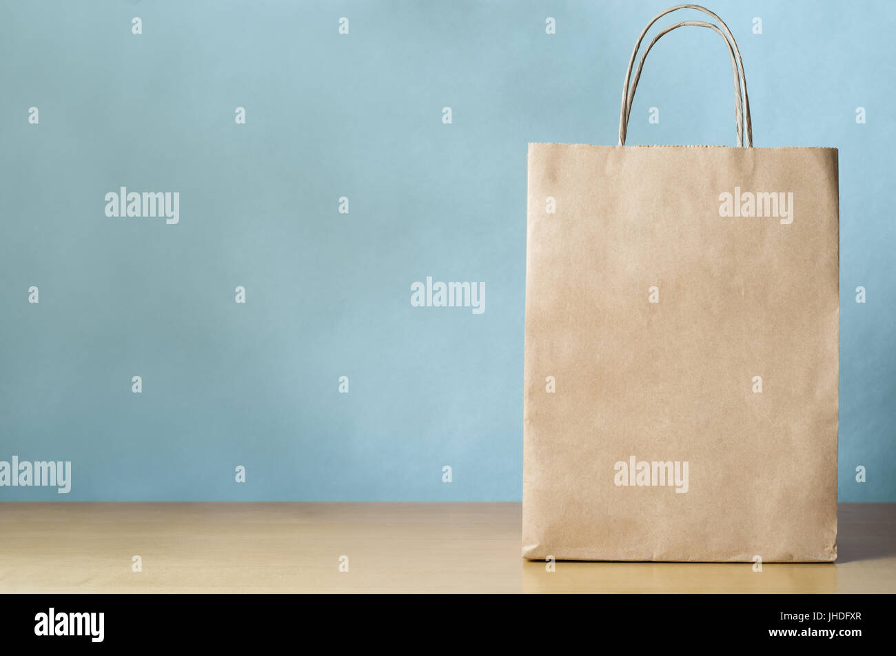Blank brown paper carrier bag with handles for shopping, facing front on right side of a light wood veneer table with pale blue wall background provid Stock Photo