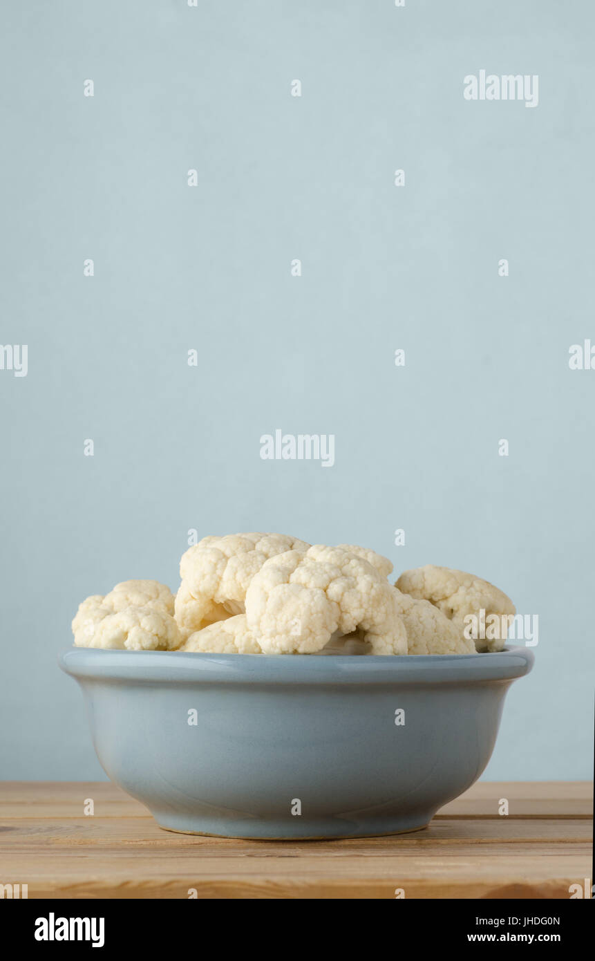 Ceramic bowl filled with raw cauliflower florets on wood plank table against light blue background with copy space above. Eye level shot. Stock Photo