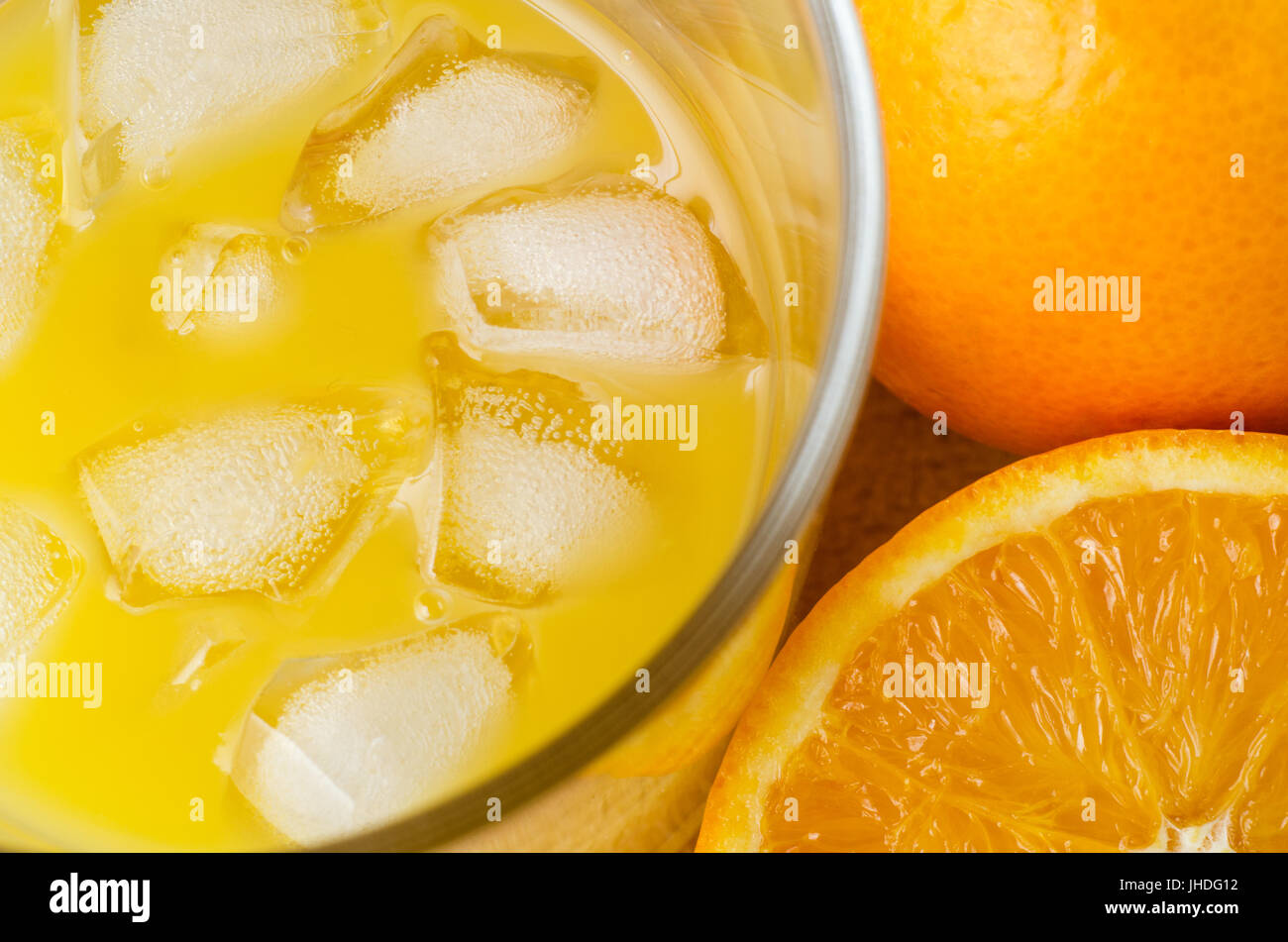 Angled overhead close up shot of fresh juice in glass with ice cubes, beside whole and half oranges on wooden table. Stock Photo