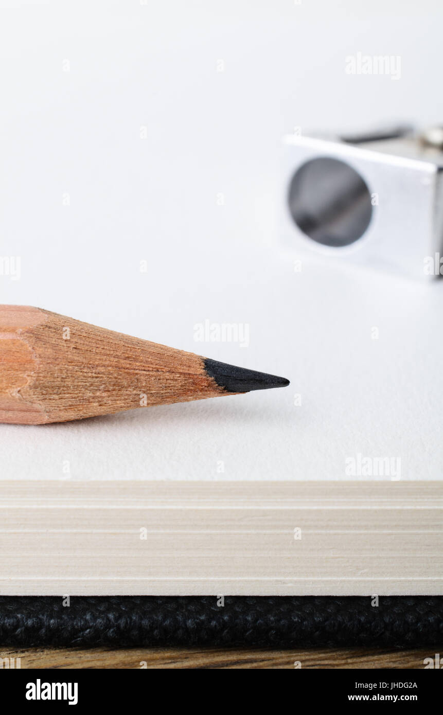 Close up of the tip of a wooden graphite drawing pencil on the edge of a blank sketch book page with sharpener in soft focus background. Stock Photo