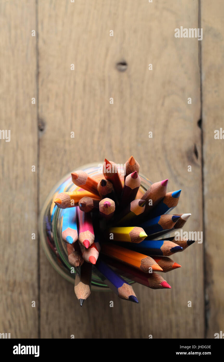 Overhead shot of art pencils in many colours, grouped in a glass jar on wooden planked table with copy space above. Stock Photo