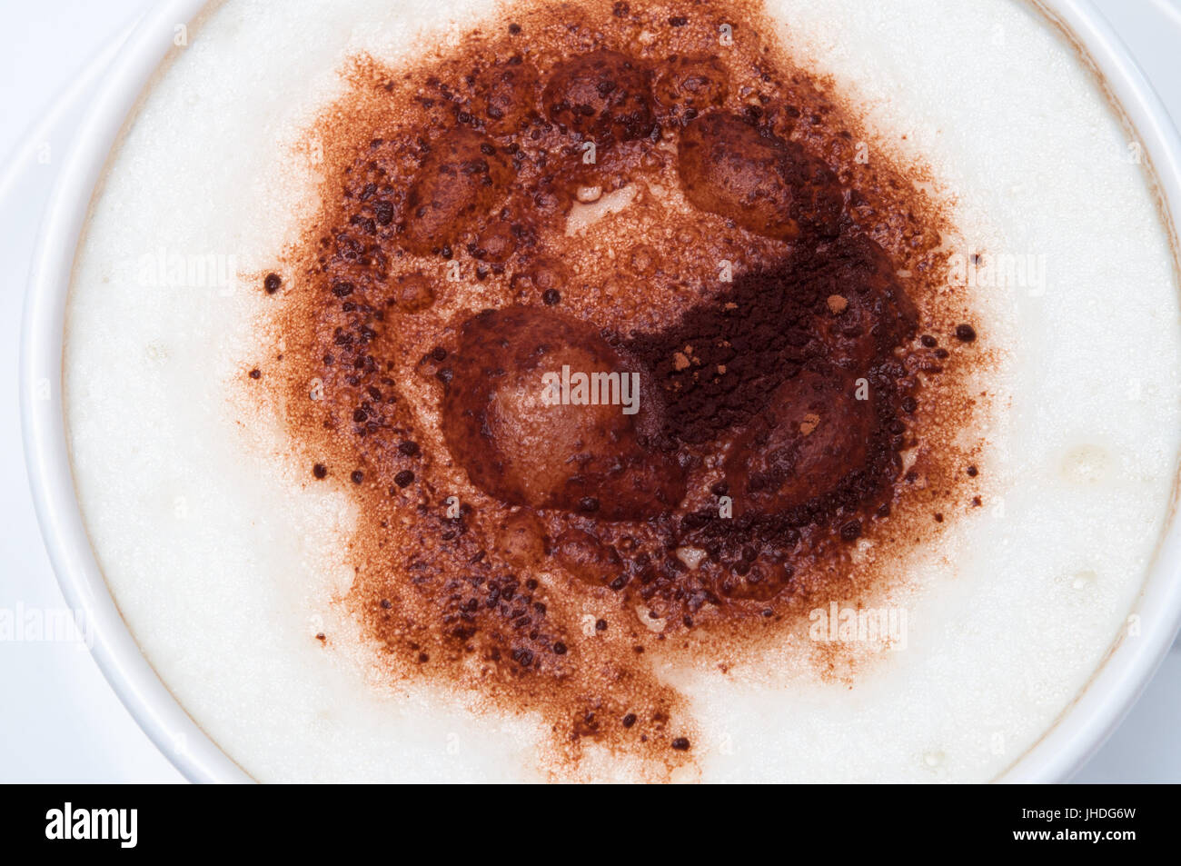 Overhead close up of milky foam and cocoa sprinkles topping a latte or cappuccino coffee in white cup with saucer. Stock Photo