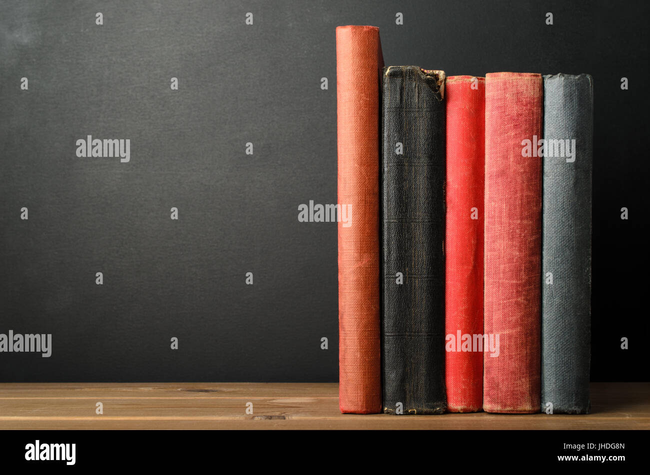 A row of upright books with blank spines at eye level on wood planked desk with black chalkboard background; providing copy space to left. Stock Photo