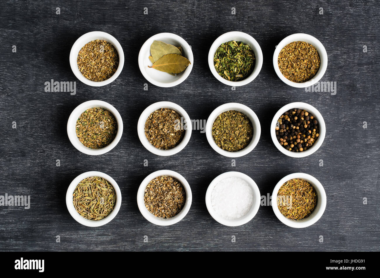 Overhead shot of rows of dried herbs, salt and peppercorns in small bowls on a dusty, textured chalkboard surface. Stock Photo