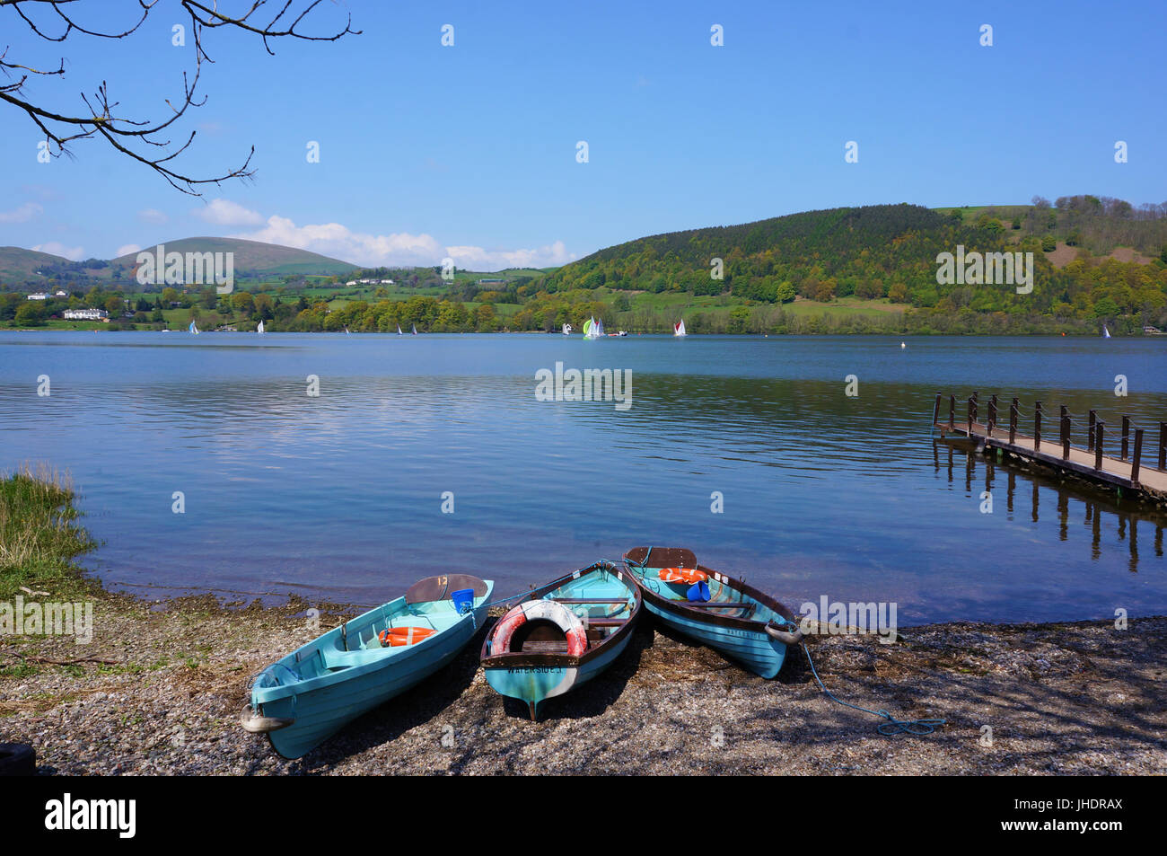 Ullswater lake on sunny day with clear blue sky.  Three rowing boats on lakeside next to small pier. View over hills, Lake District England. Stock Photo