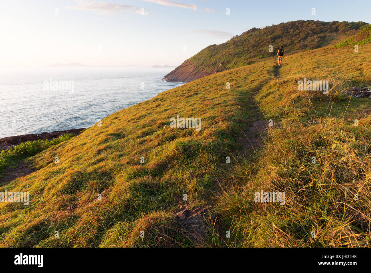 A hiker walks on a grassy coastal trail as bright morning light warms the coastline that surrounds. Stock Photo
