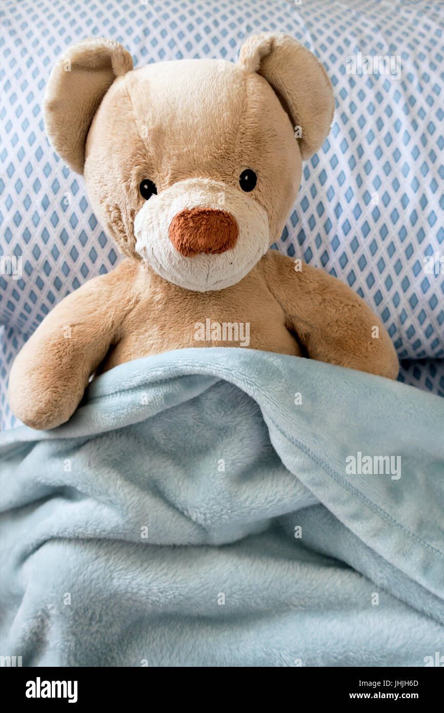 A tan teddy bear sitting in bed with a blue checkered pillow and blue blanket. Stock Photo