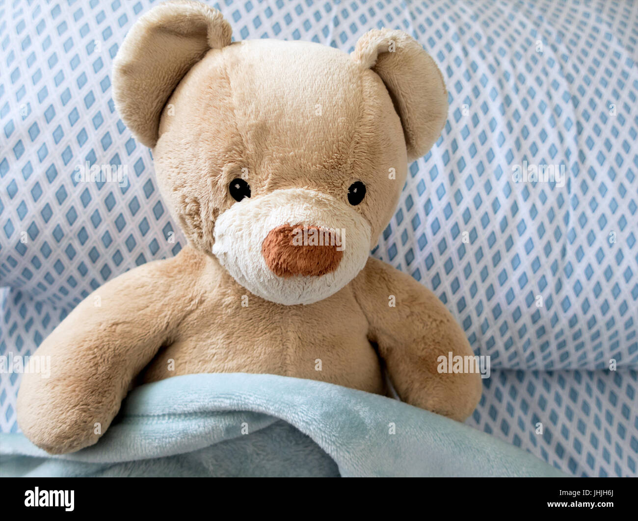 A tan teddy bear sitting in bed with a blue checkered pillow and blue blanket. Stock Photo