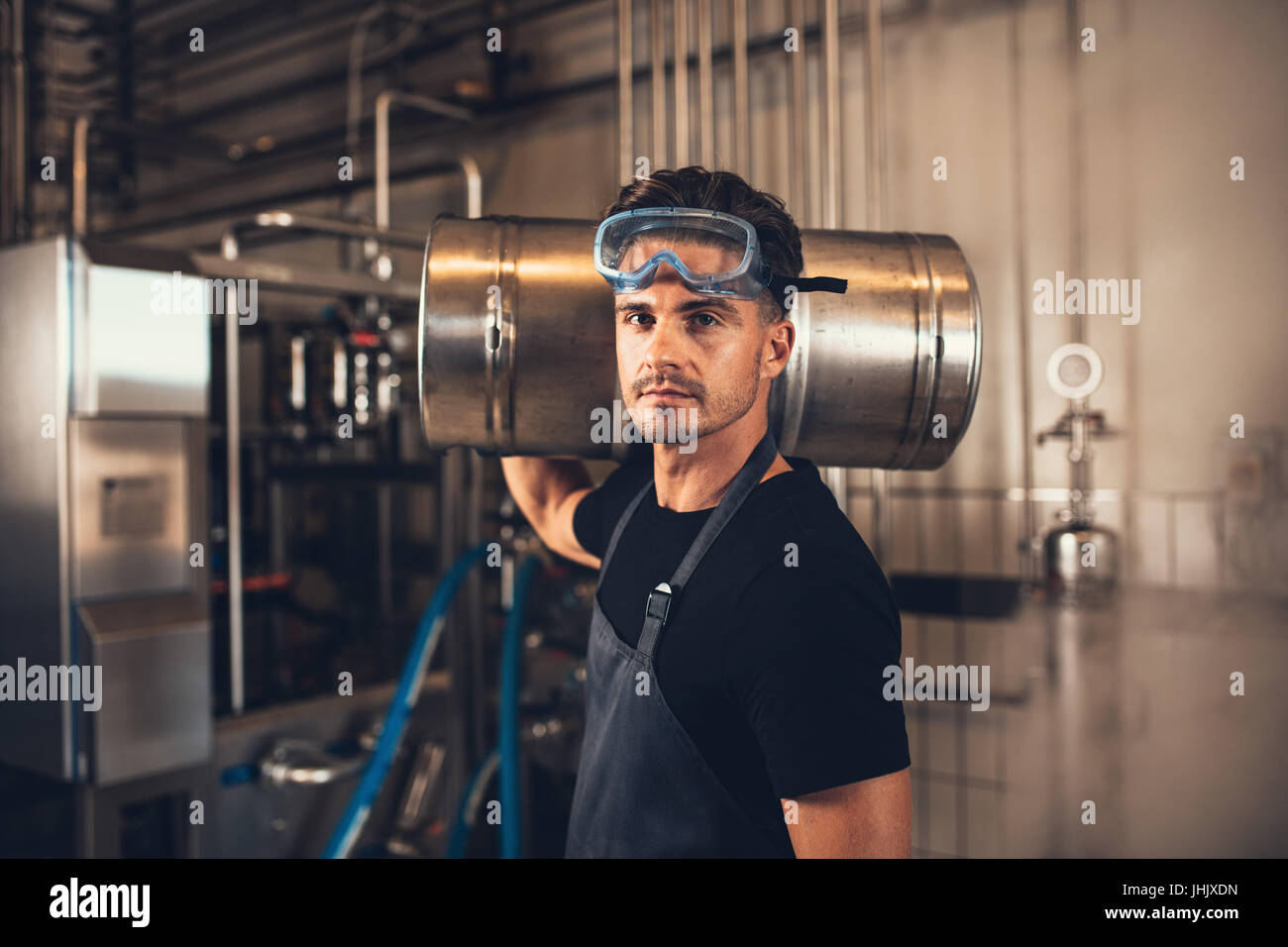 Portrait of male brewer with keg at brewery factory. Young man holding a metal beer container on shoulder at warehouse. Stock Photo