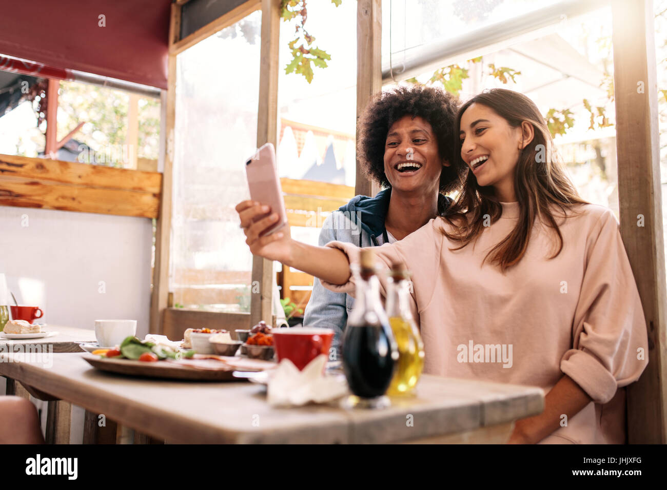 Cheerful woman taking selfie with her friend at cafe. Couple having fun using smartphone at coffee shop Stock Photo