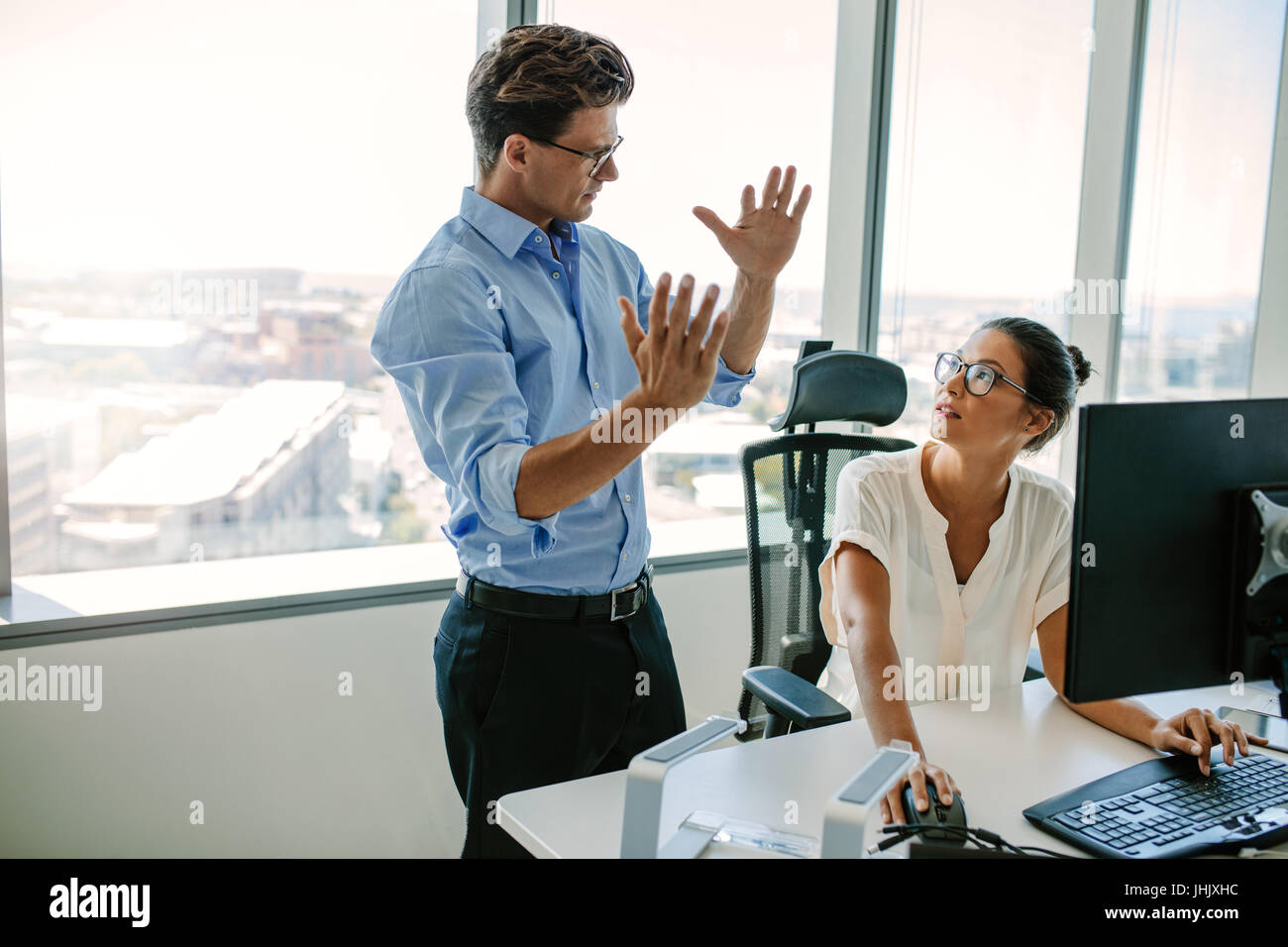 Mature man explaining new business strategy to asian female colleague sitting at her desk. Two business professionals working together in office. Stock Photo