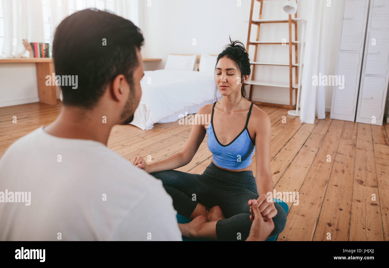 Beautiful young woman doing yoga at home with her instructor. Young couple sitting together holding hands and practising yoga. Stock Photo