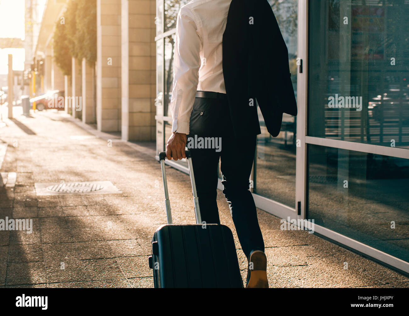 Rear view of young businessman walking outside public transport building with suitcase. Business traveler pulling suitcase on city street. Stock Photo