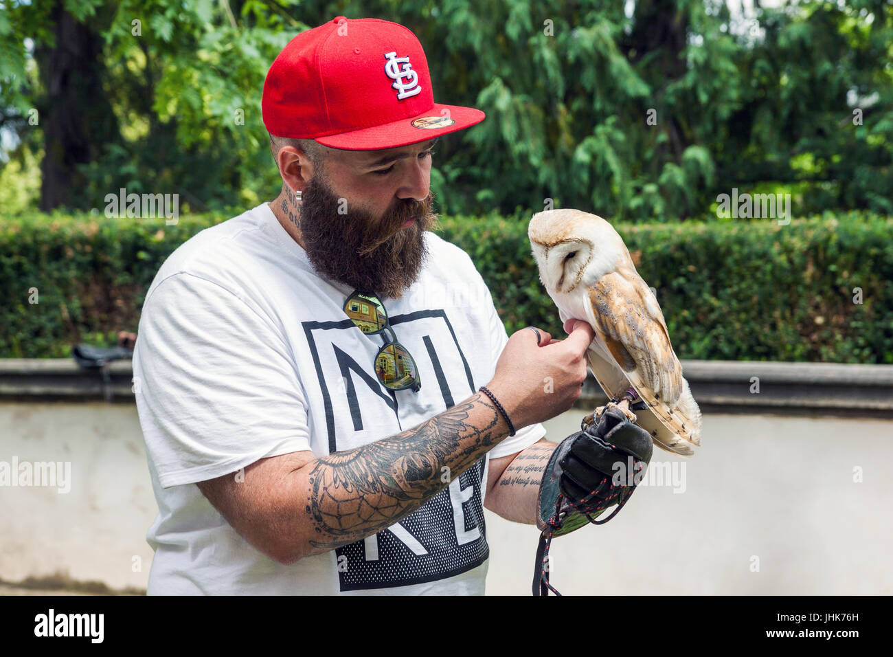 Bearded caucasian hipster in red full cap with tattoo on his arms holds an owl in hand in summer time on the s Stock Photo