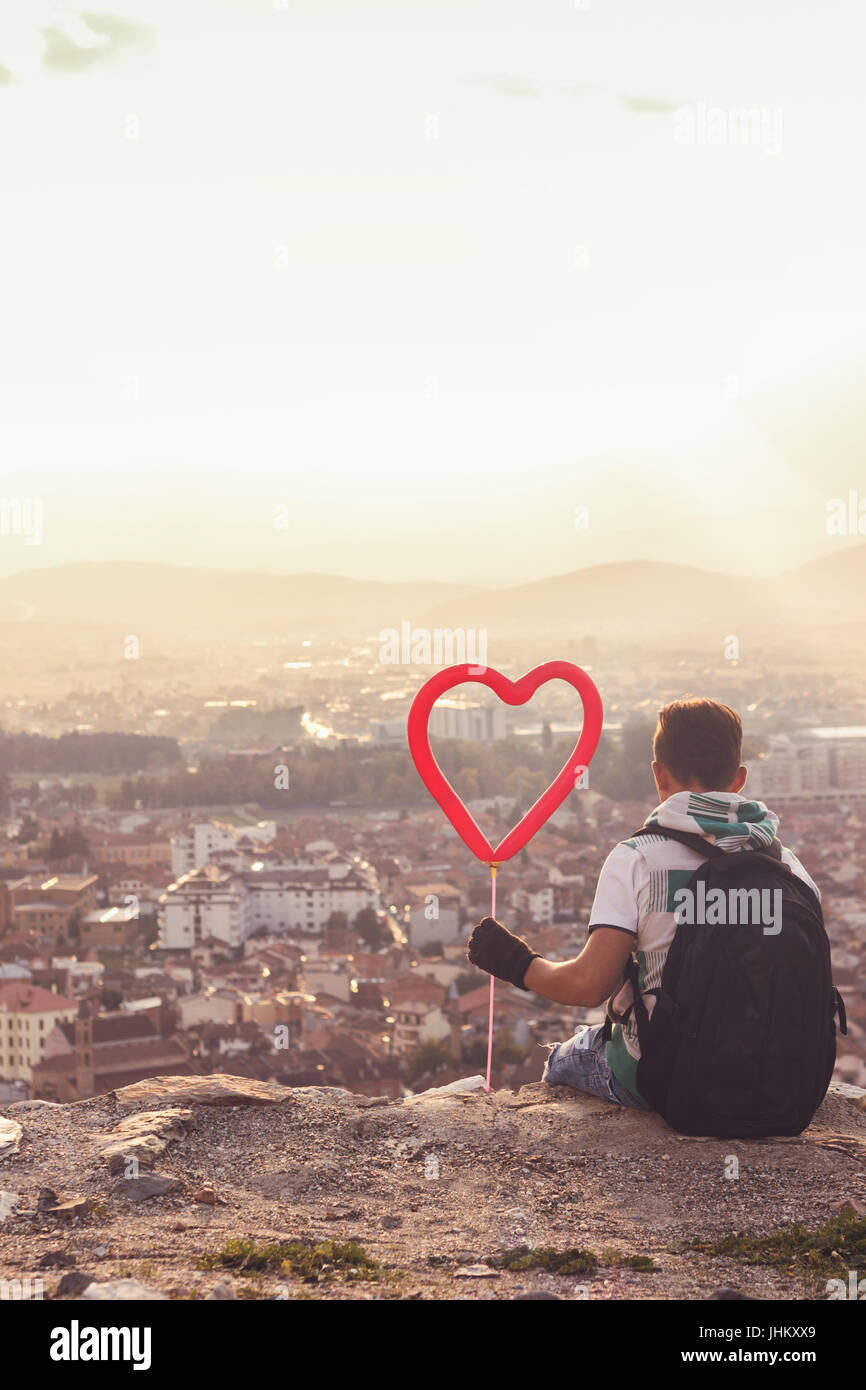 young boy with backpack and heart balloon sitting on hill in front of Prizren, Kosovo. He looking to the city from high viewpoint Stock Photo