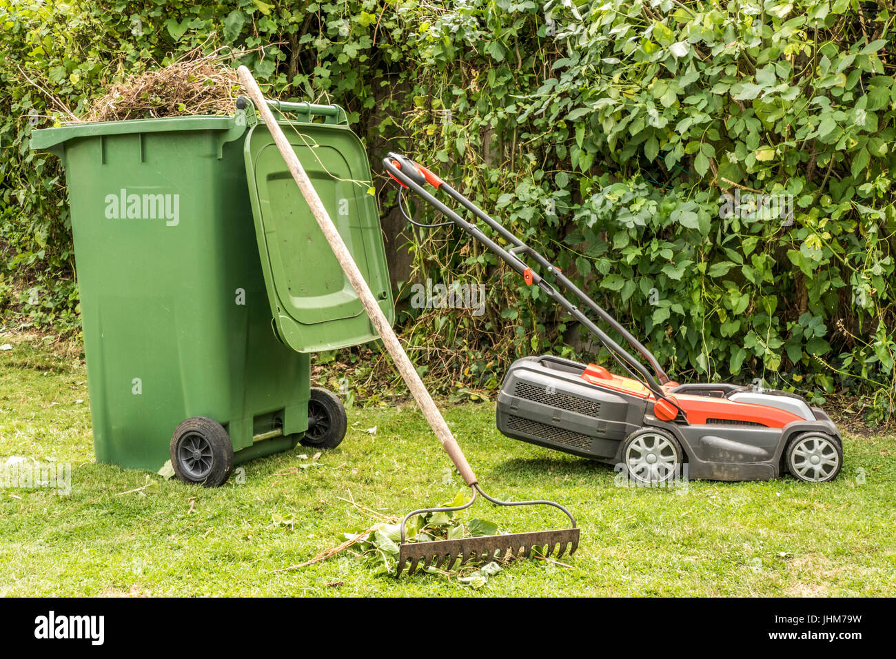 A lawn mower, a wheely bin full of cuttings and a rake, in a garden in England, UK. Stock Photo