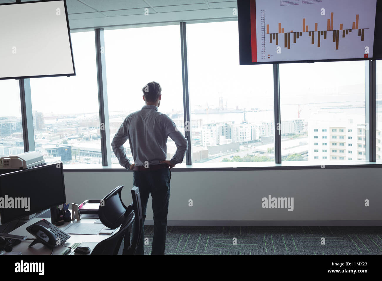 Rear view of businessman standing with hands on hip against window at office Stock Photo