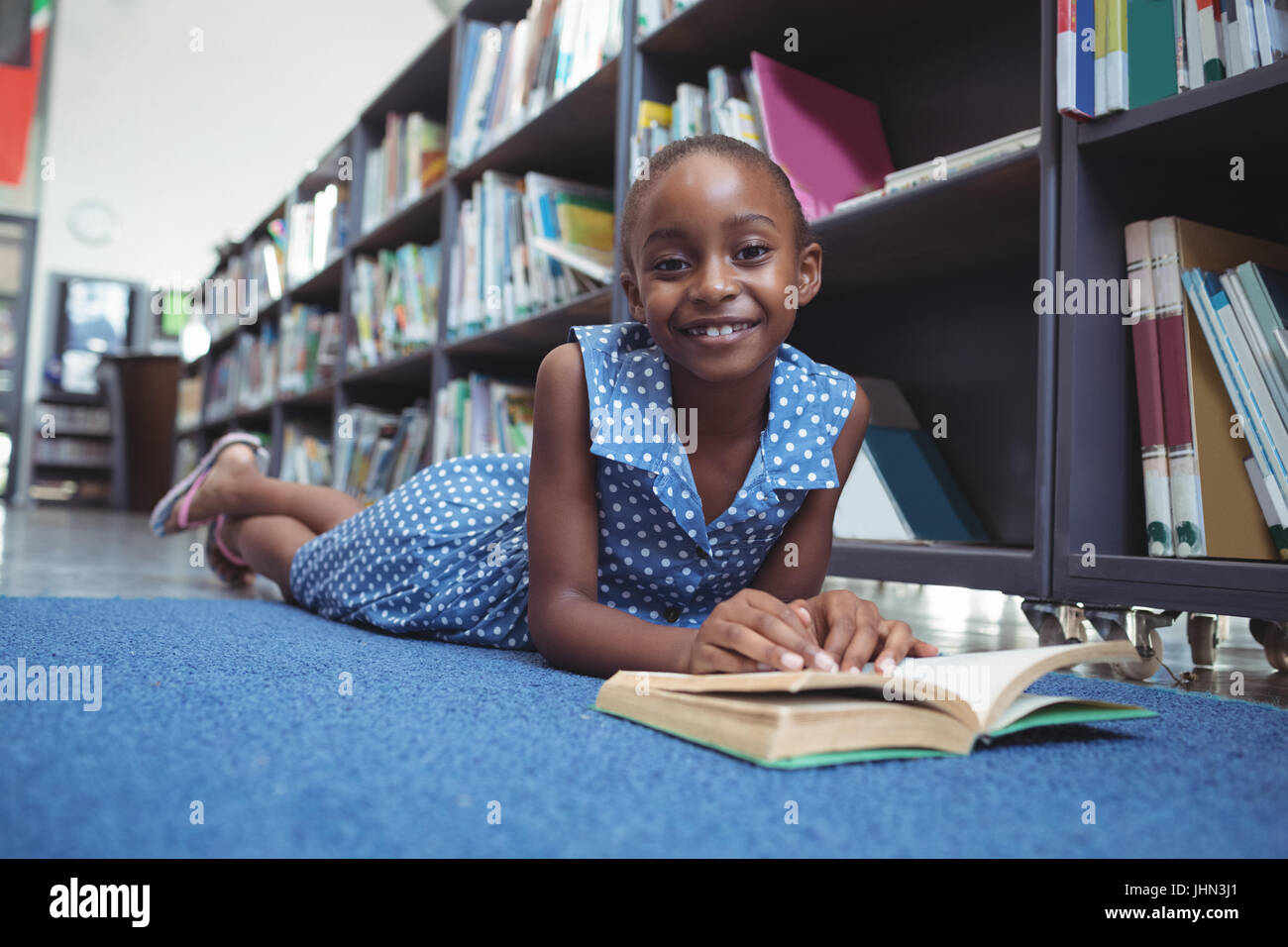 Portrait of smiling girl with book lying by shelf in library Stock ...