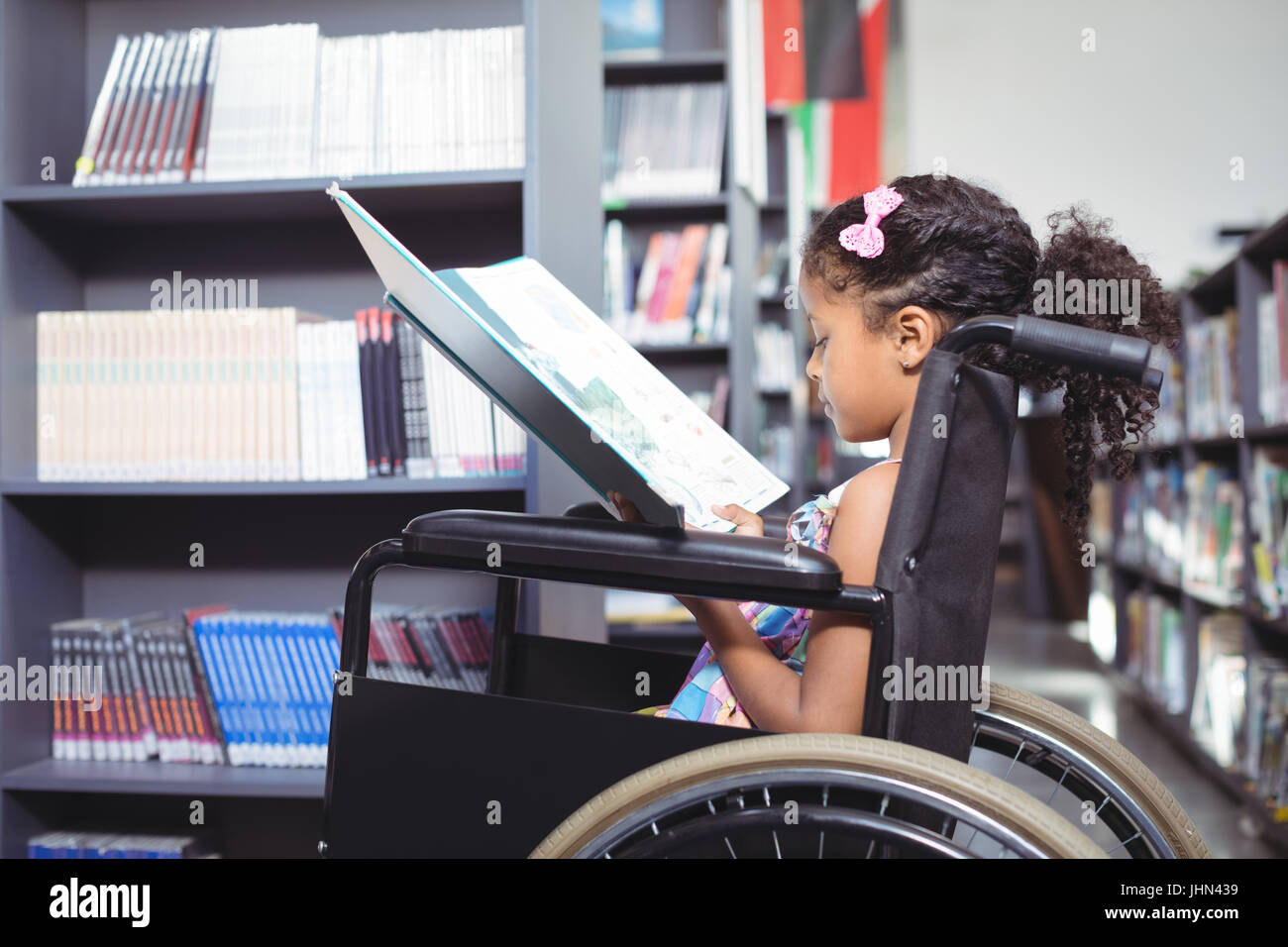 Side view of girl reading book while sitting on wheelchair in library ...