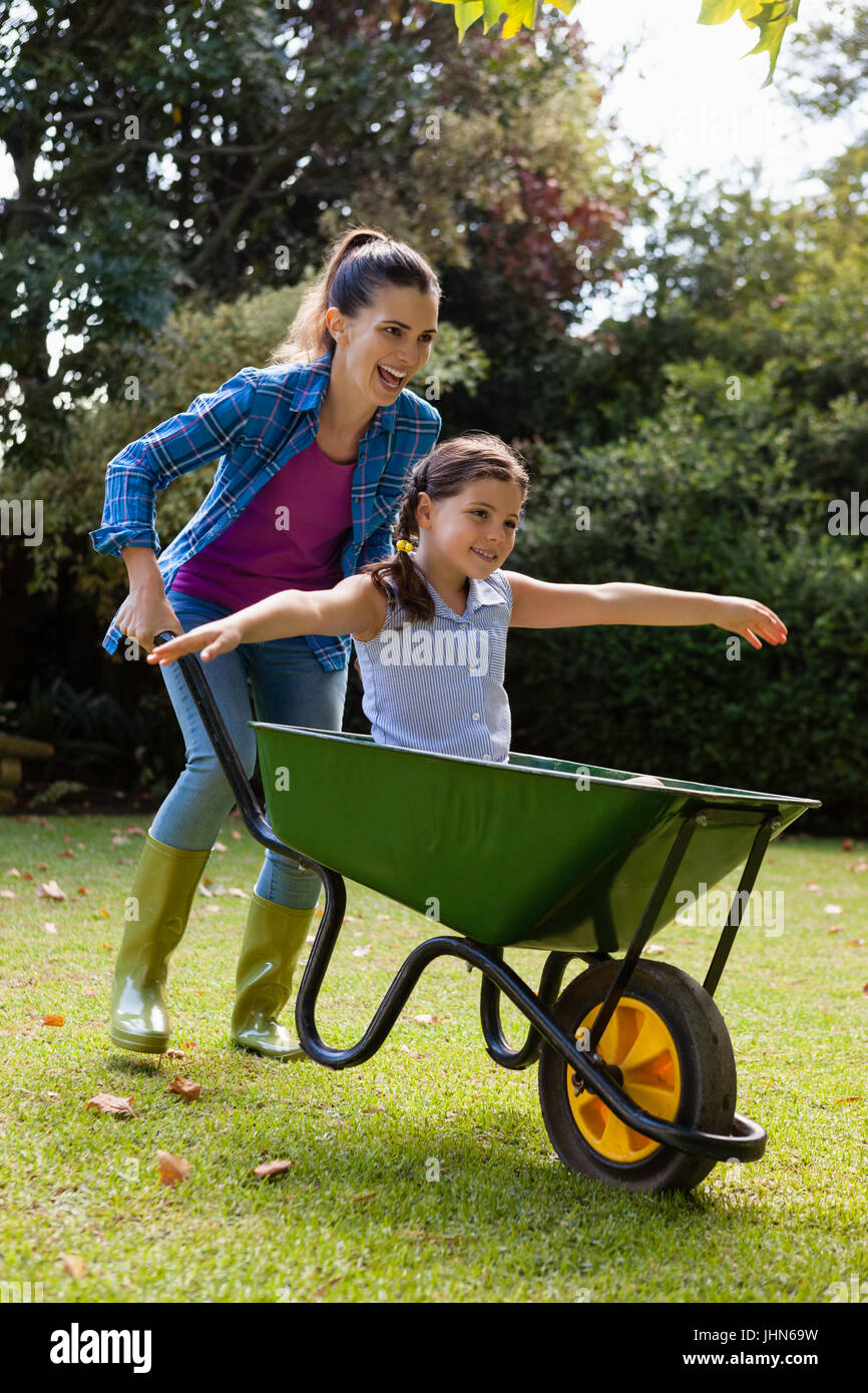 Cheerful mother pushing daughter sitting in wheelbarrow at backyard ...