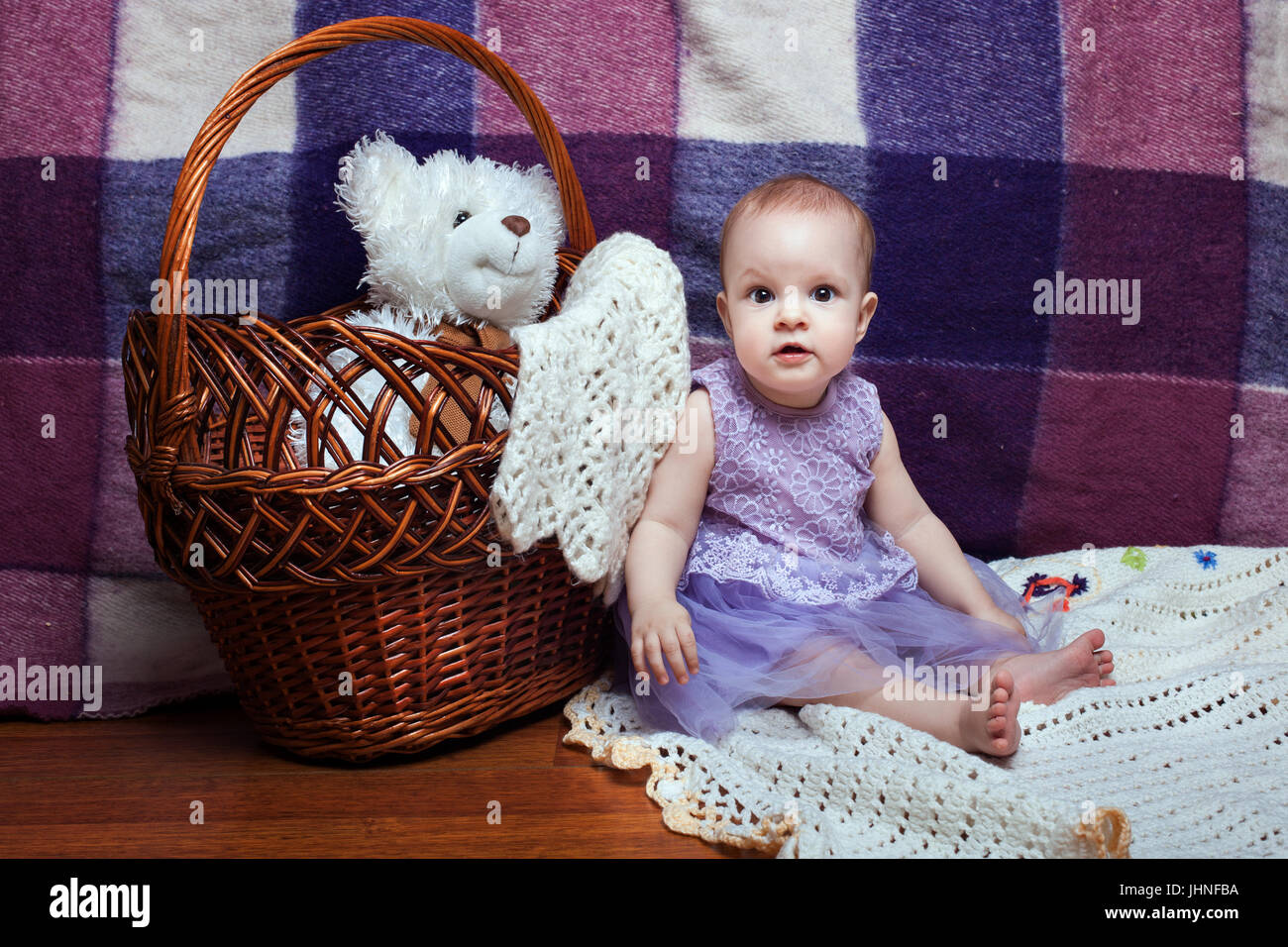 Adorable baby girl in a lilac dress sitting near wicker basket Stock Photo