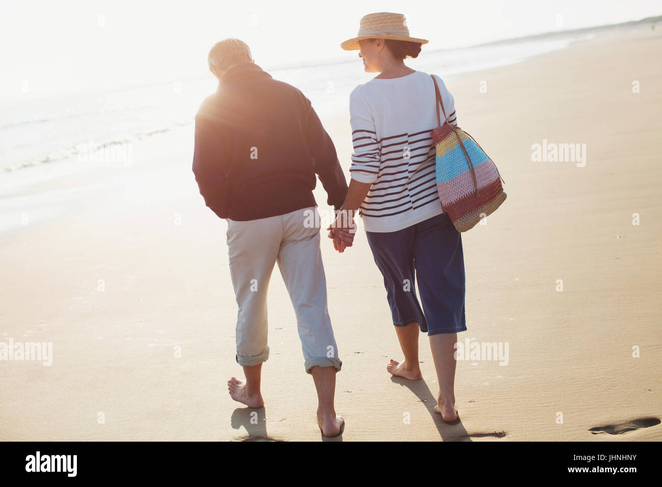 Barefoot mature couple holding hands and walking on sunny beach Stock Photo