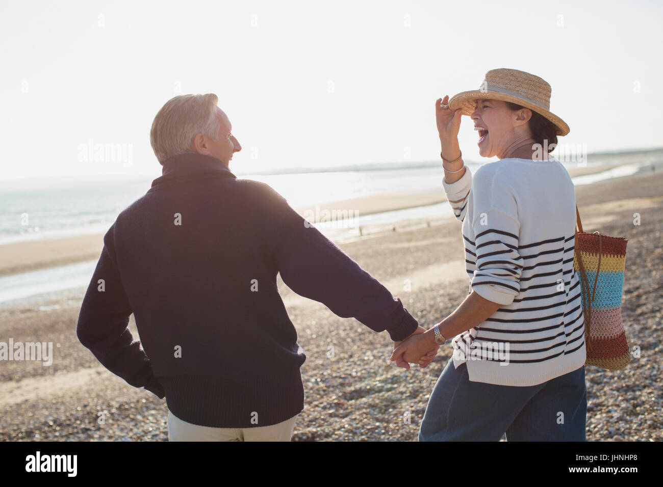 Laughing mature couple holding hands and walking on sunny beach Stock Photo