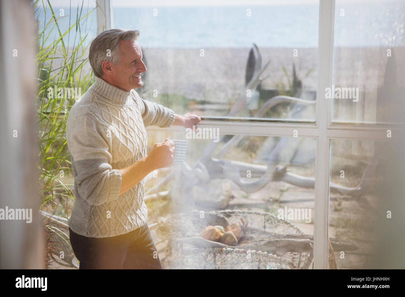 Mature man in turtleneck sweater drinking coffee in sunny beach house sun porch Stock Photo