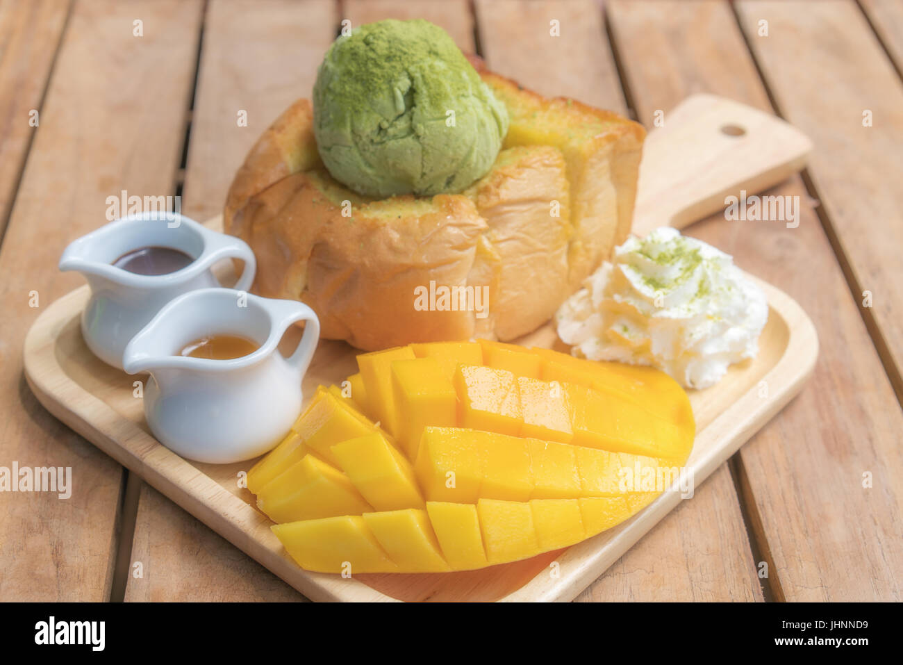 Honey toast green tea ice-cream with ripe barracuda mango and whipping cream in wooden plate on wooden table. Stock Photo