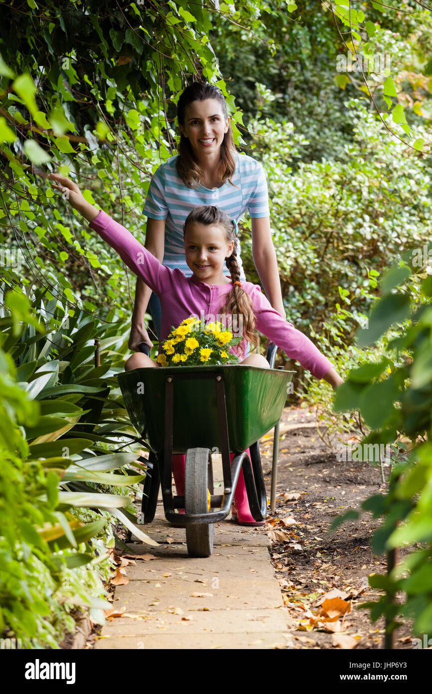 Woman pushing girl sitting with outstretched in wheelbarrow on footpath ...