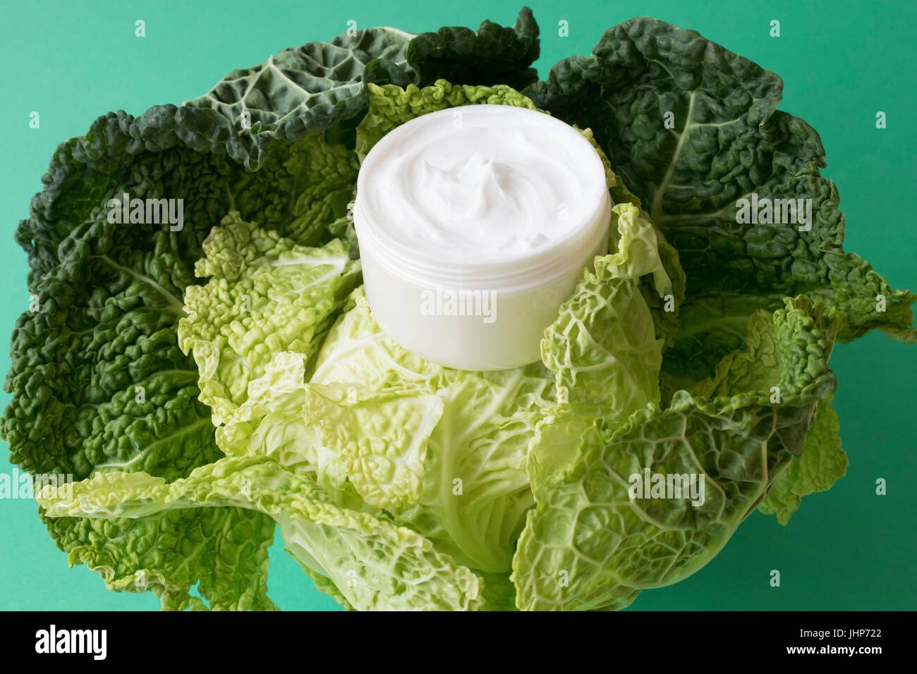 Pot of moisturizing cream and cabbage leaves against a green background. Conceptual image of natural ingredients in cosmetics. Stock Photo