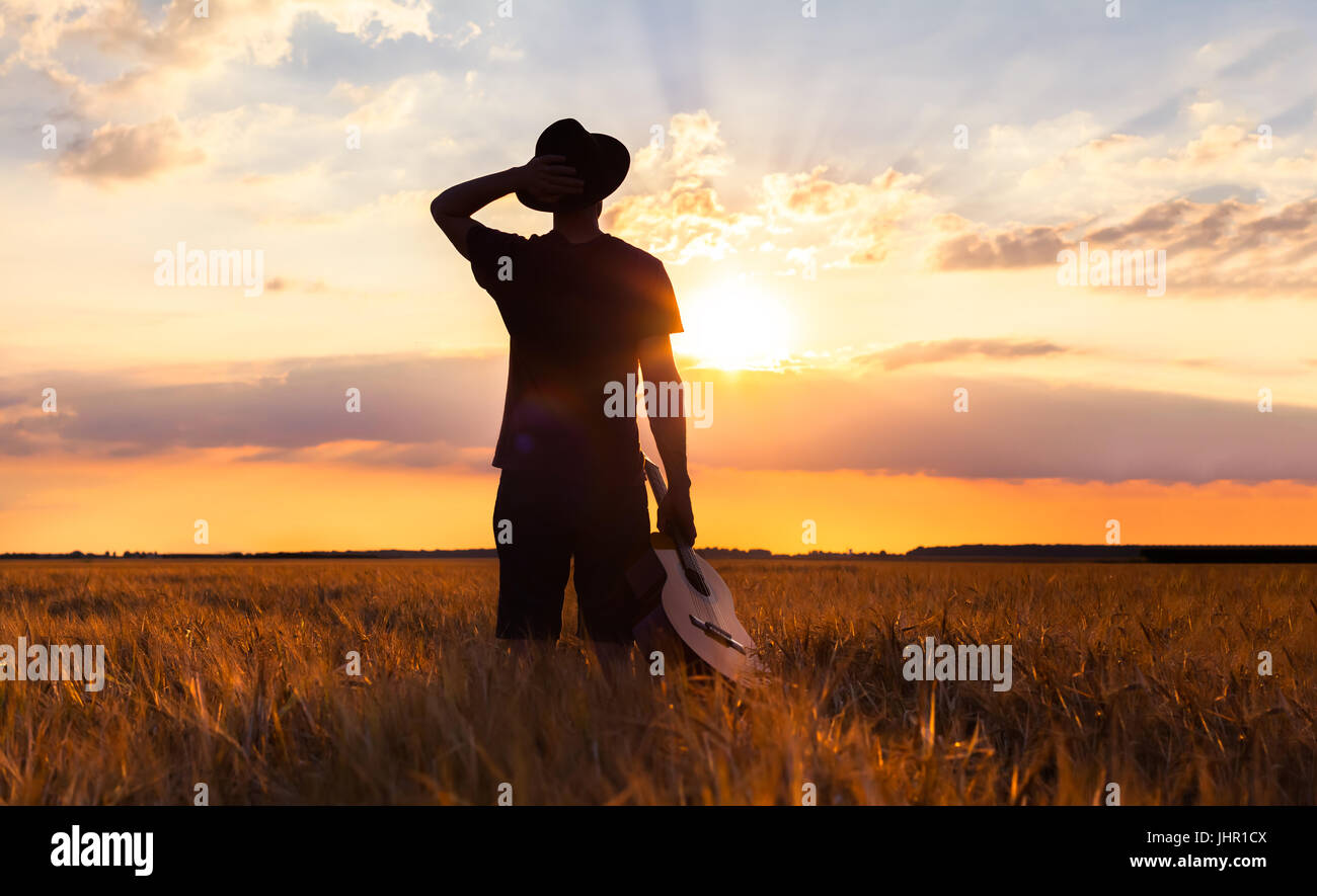 Person standing with his guitar in the warm summer fields looking at sunset for musical inspiration Stock Photo
