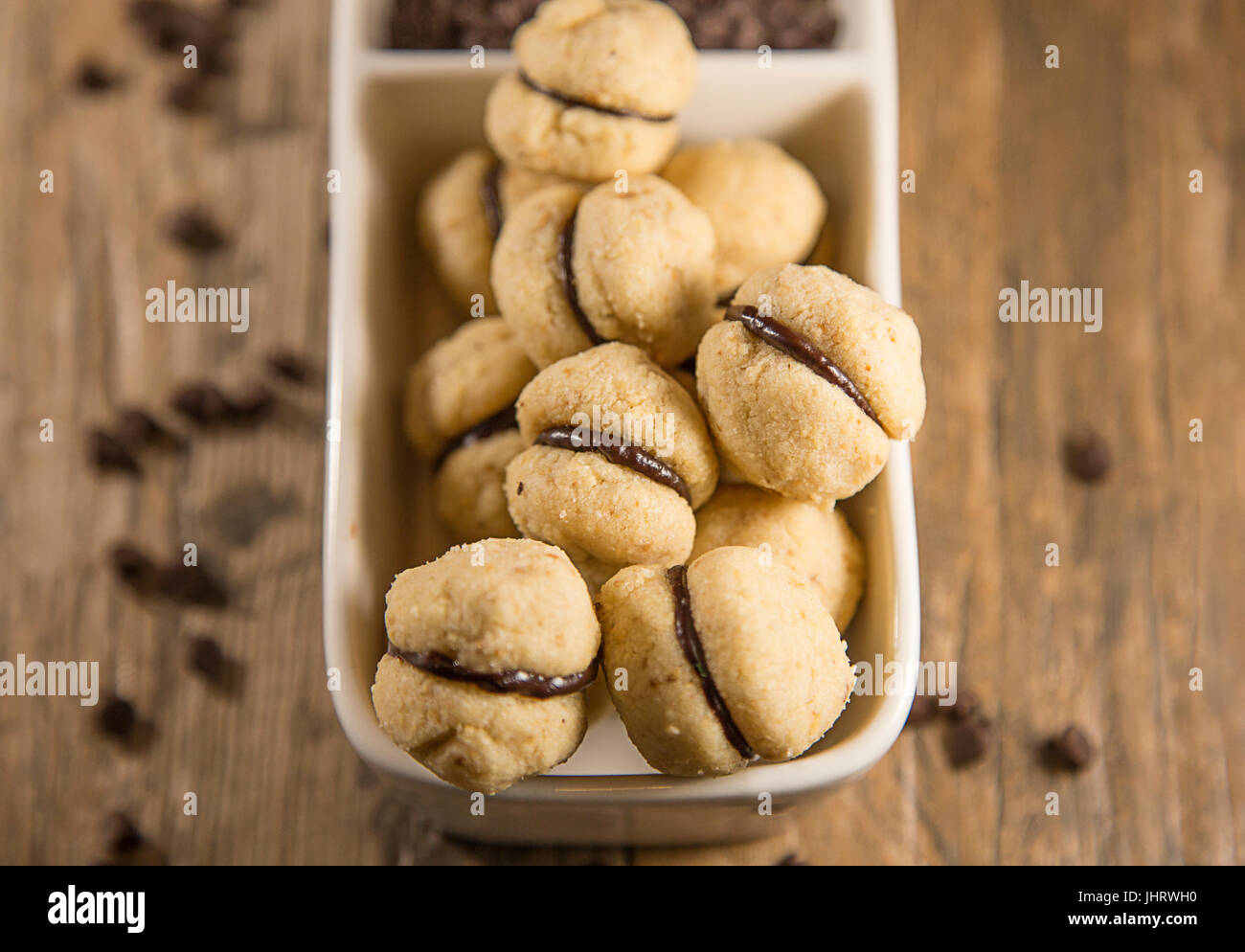 Bite-size round mini cookies with hazelnut filling, displayed in a white serving dish, with chocolate chips. Wooden background. Stock Photo