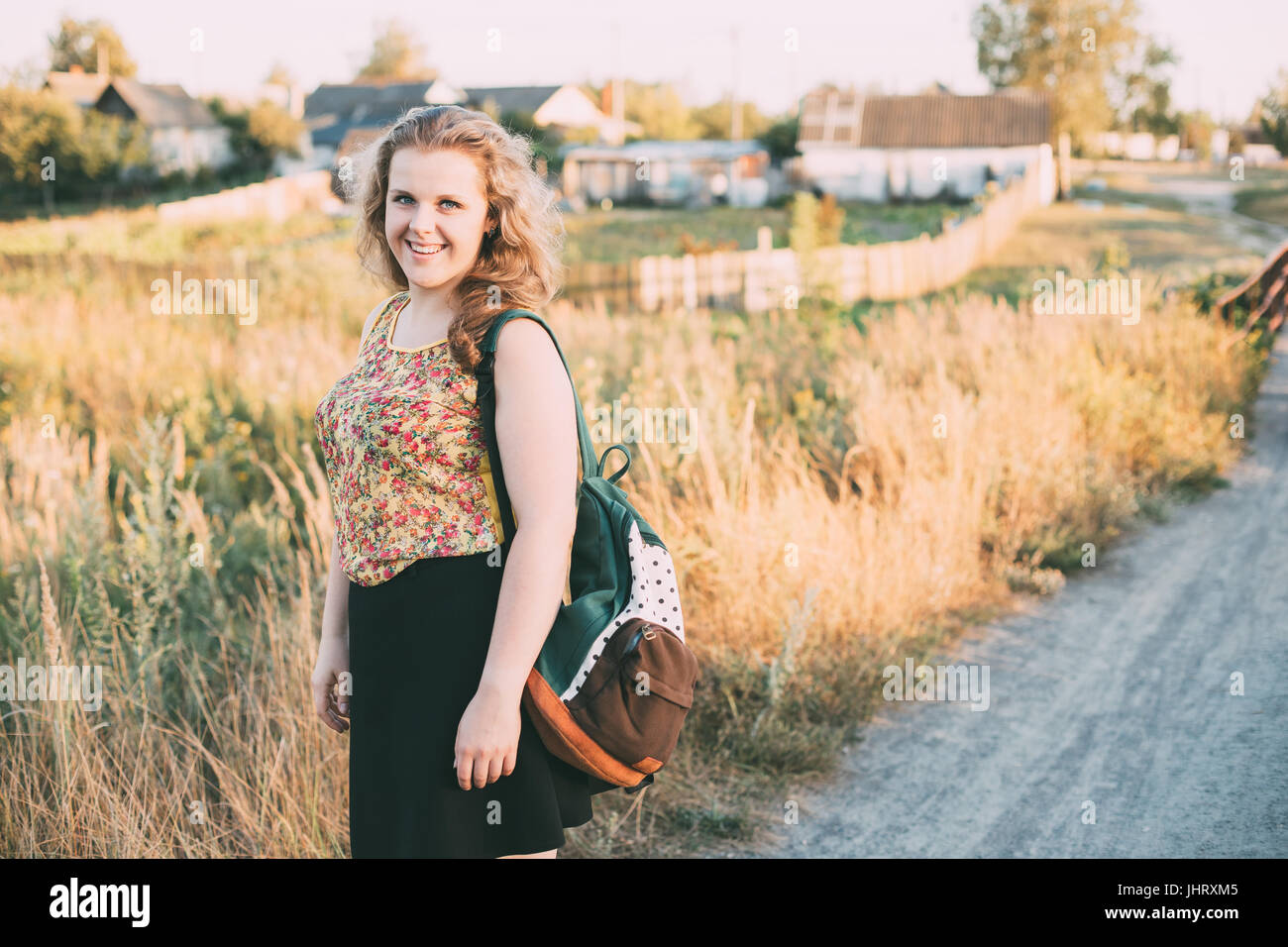 Portrait Of Young Pretty Plus Size Caucasian Happy Girl Woman With Blue Eyes, Wavy Brown Long Hair And Backpack In Summer Meadow Stock Photo