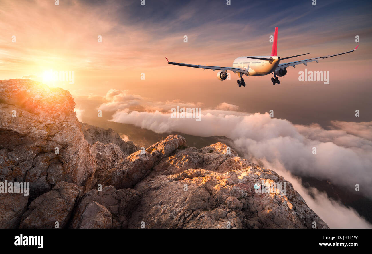 Airplane is flying over clouds at sunset. Landscape with white passenger airplane, mountains, sea and orange sky with sun in summer. Passenger aircraf Stock Photo