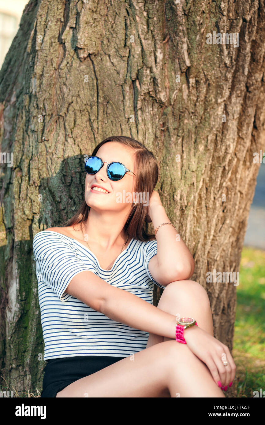 young beautiful happy woman in round sunglasses sitting on the grass near a tree in the park Stock Photo
